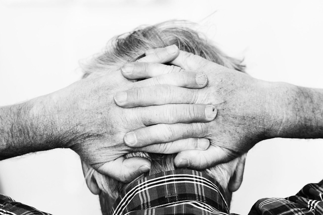 An older man faces away from the camera with his hands clasped behind his head as if he has a headache