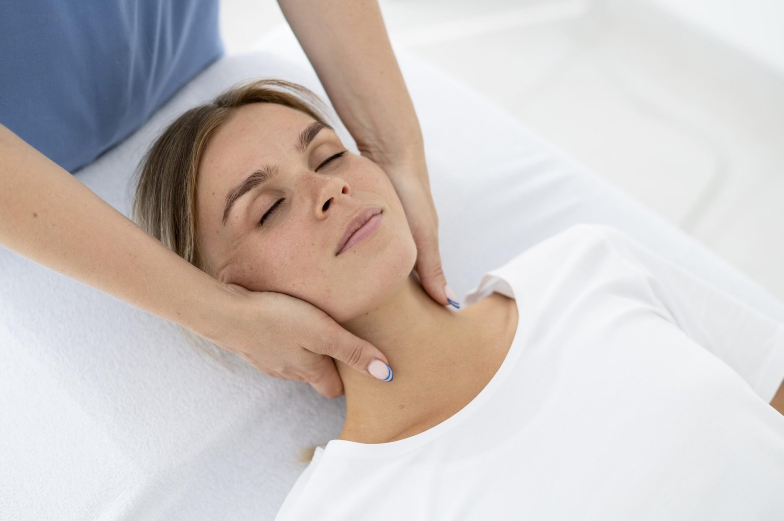 A woman lies face up on a plinth with her eyes closed while a therapist is standing at the head of the bed holding on to both sides of her head