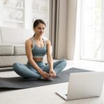 A woman sits on an exercise mat in a butterfly position in front of a laptop computer in a brightly lit lounge room
