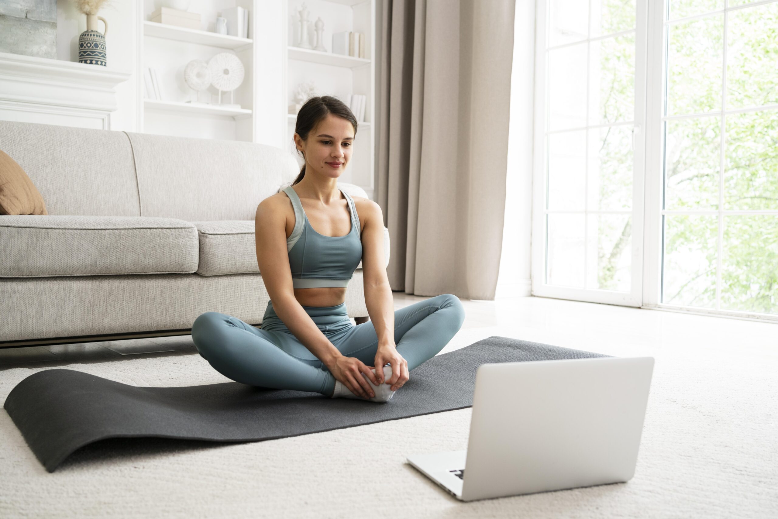 A woman sits on an exercise mat in a butterfly position in front of a laptop computer in a brightly lit lounge room